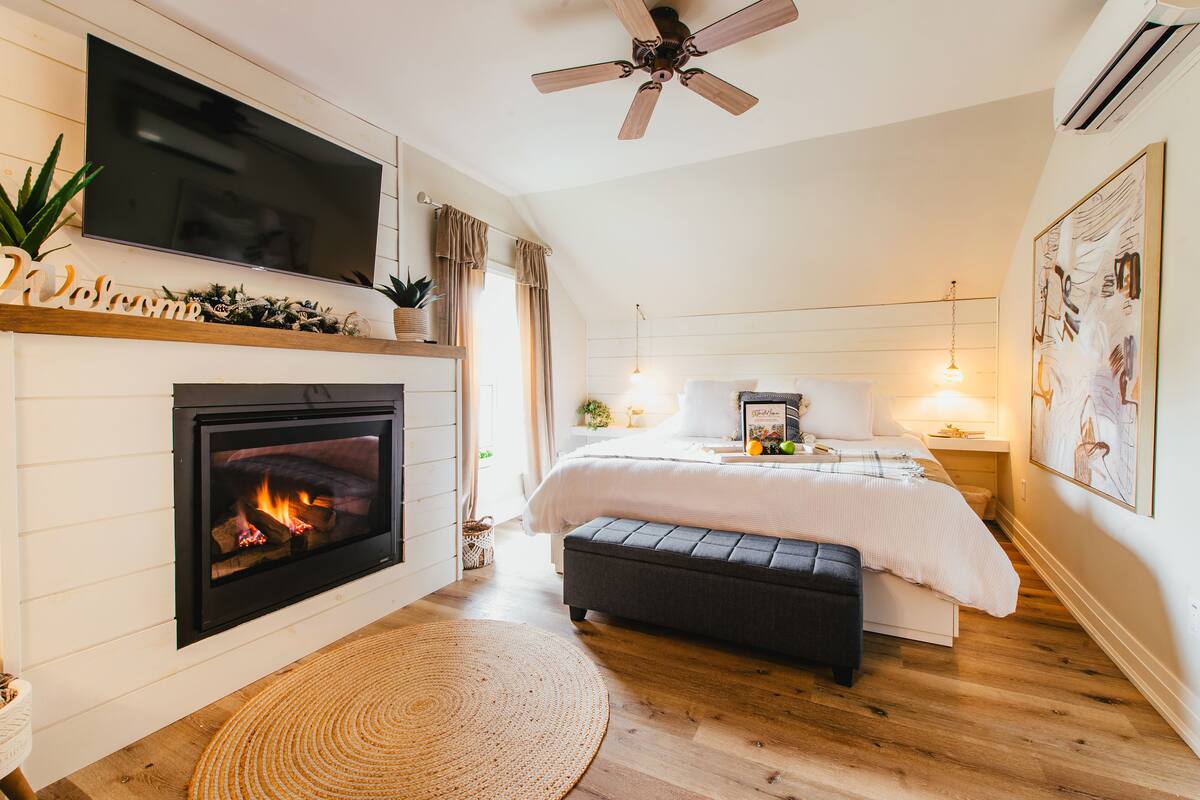 Une chambre avec une cheminée, un lit, un ventilateur de plafond et un téléviseur fixé au mur.