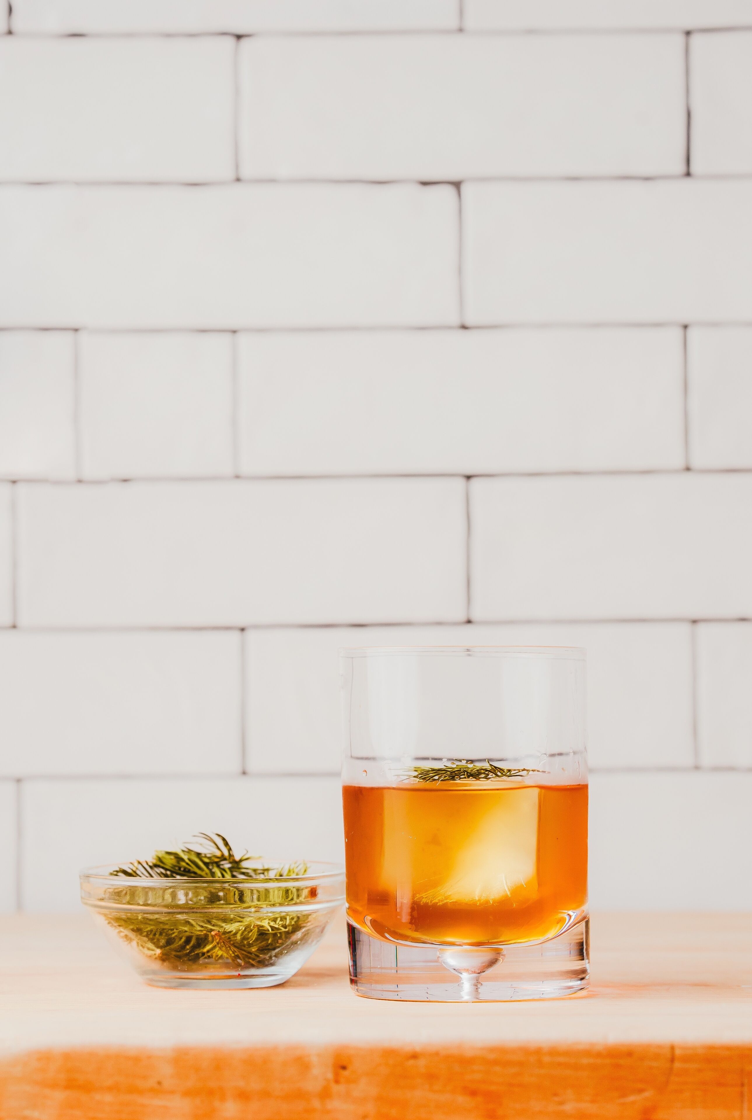 A clear glass of amber liquid with a sprig of rosemary, placed on a wooden surface against a white brick wall.