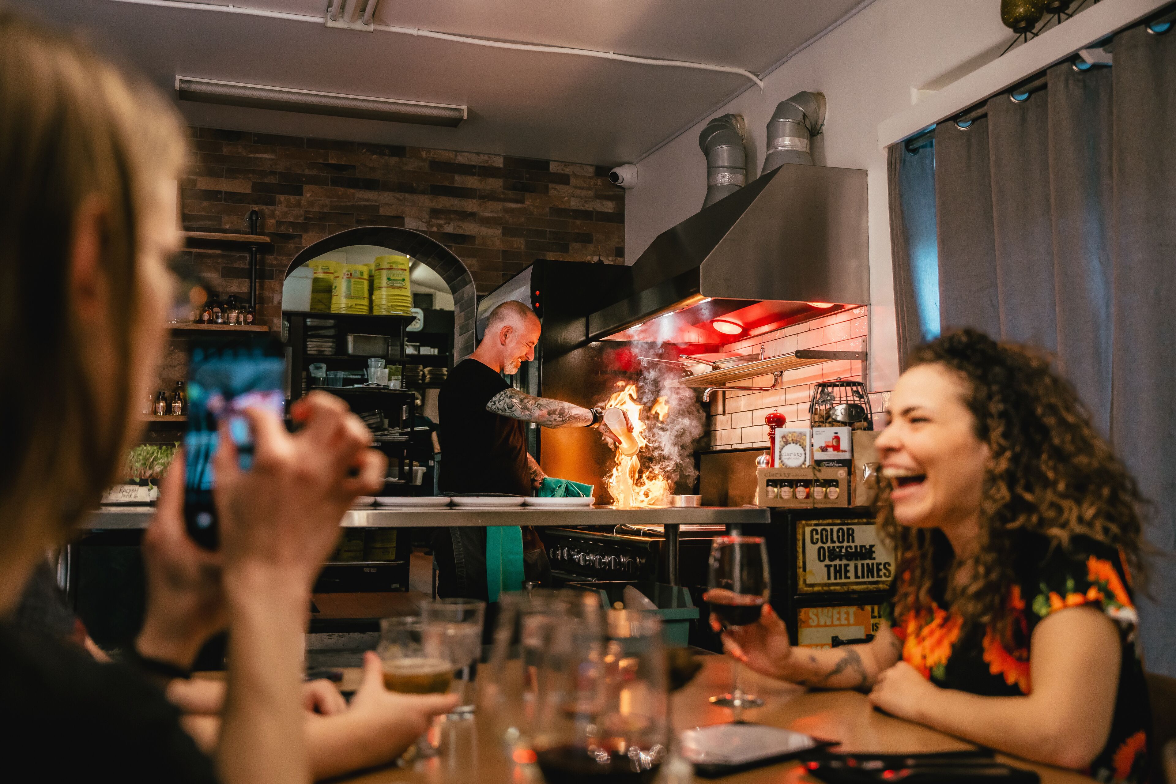 Un bar con pared de ladrillo, una persona cocinando y vasos de bebidas sobre la mesa.