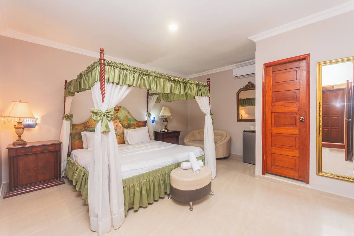 A four-poster bed with a canopy, a bedside table with a lamp, a chair, and a small ottoman.