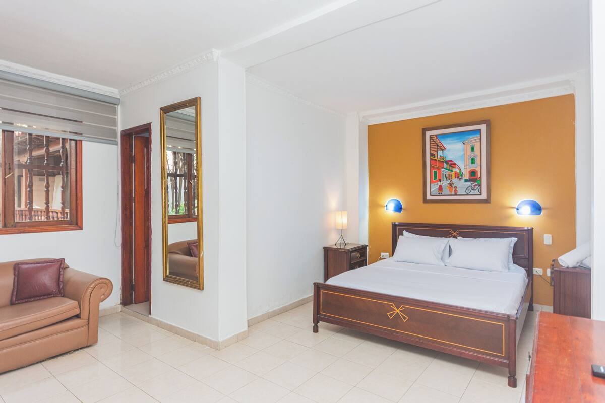 A hotel room with a bed, a nightstand, a lamp, a mirror, a sofa, and a painting on the wall.