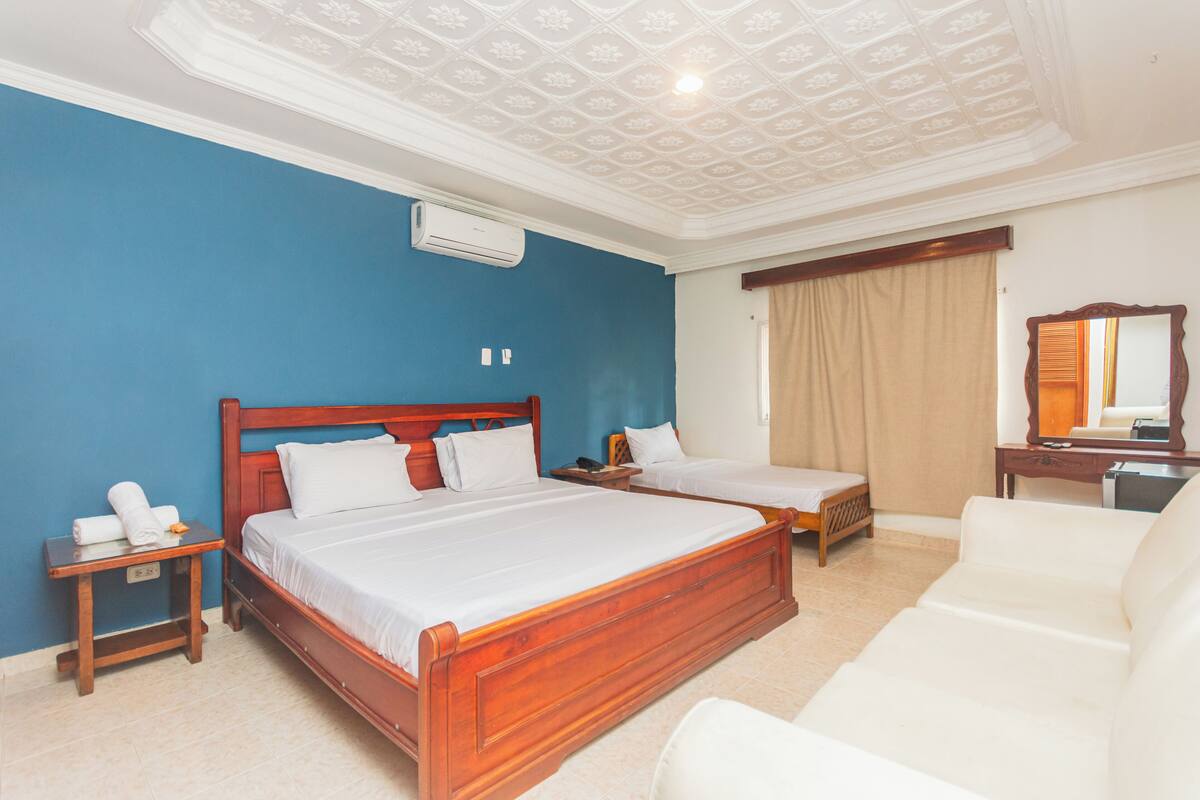 A hotel room with a large bed, two smaller beds, a sofa, a mirror, and a ceiling with a decorative pattern.