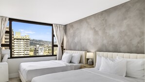 A hotel room with two beds, a large window with a view of buildings, a white headboard, and a bedside table with a lamp.