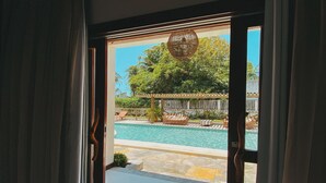 A view through a window with curtains, revealing a pool area with a pergola, lounge chairs, and a wooden fence. - Pousada Velho Chico (Amontada)