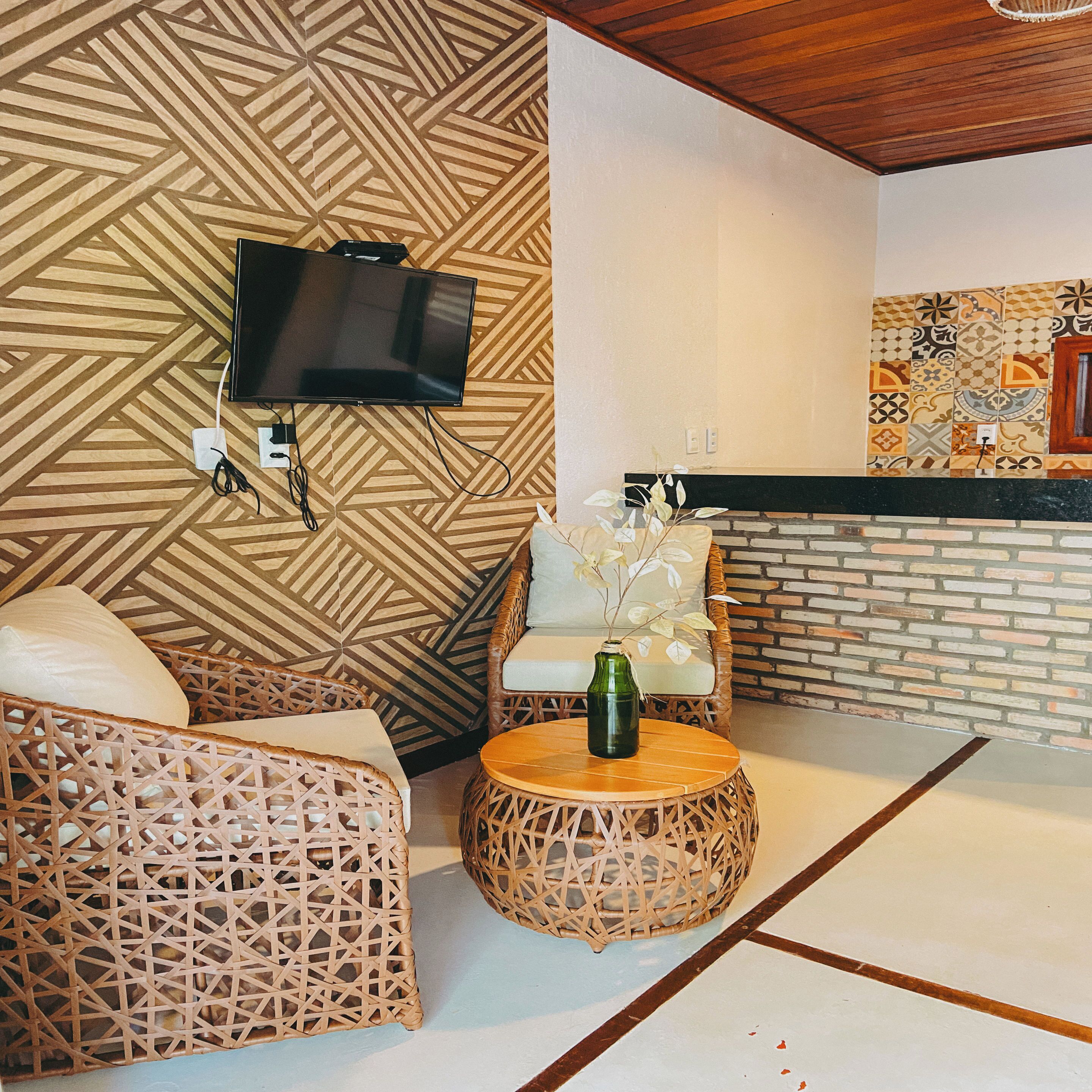 Una habitación con televisión montada en una pared de diseño geométrico, muebles de mimbre, una mesa redonda de madera con un jarrón y una chimenea de ladrillo.