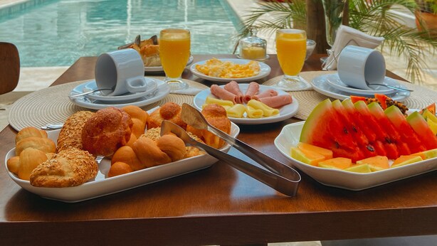 A breakfast table with a pool view, including bread, fruit, and juice. - Pousada Velho Chico (Amontada)