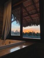 Un baño con bañera, ventana con vista a palmeras y un atardecer, y techo de paja.