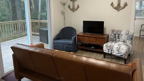 Un salon avec un canapé en cuir marron, un fauteuil bleu, une table basse en bois et un téléviseur posé sur un meuble en bois. Deux éléments décoratifs en forme d’ancre ornent le mur, et une lampe sur pied est placée dans un coin de la pièce.
