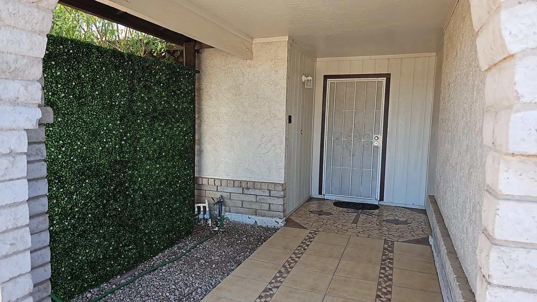 Una entrada techada con piso de baldosas, una puerta con reja de metal y un seto verde.