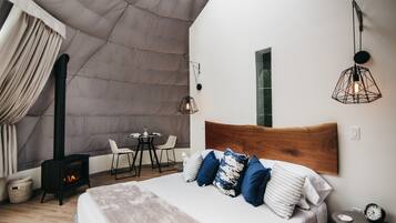 A modern bedroom with a large bed, a wooden headboard, a small table with chairs, a wall-mounted light fixture, and a fireplace.