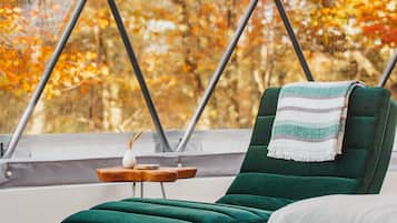 Una silla lounge verde con una toalla blanca, una pequeña mesa de madera con un florero y vistas a árboles otoñales a través de un amplio ventanal.