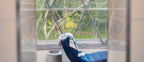 Una habitación con un ventanal grande, una silla azul y un objeto cilíndrico en el alféizar.