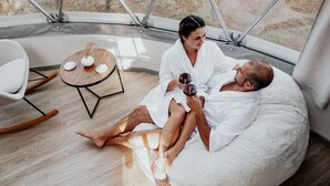 Un couple se trouve dans une pièce chaleureuse et moderne, dotée d’une grande fenêtre, d’une table ronde en bois et d’un coin salon confortable.
