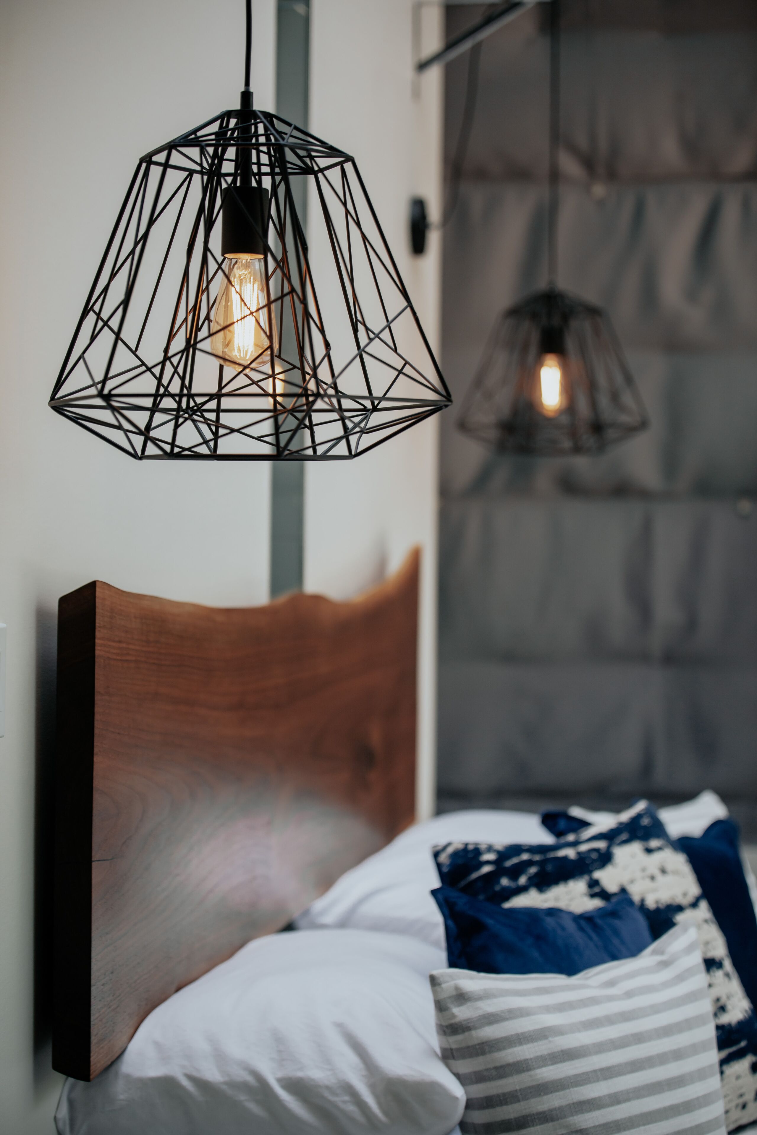 A modern bedroom with a wooden headboard, a bed with white and striped pillows, and two geometric pendant lights.