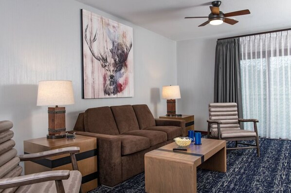 Una habitación de hotel moderna con un sofá marrón, dos mesitas de noche con lámparas, una mesa de centro de madera, una silla azul y un ventilador de techo.
