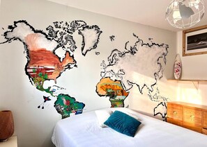 Ein Schlafzimmer mit Wandgemälde einer Weltkarte, einem Bett mit blauem Kissen, einer Holzkommode und einer Pendelleuchte.