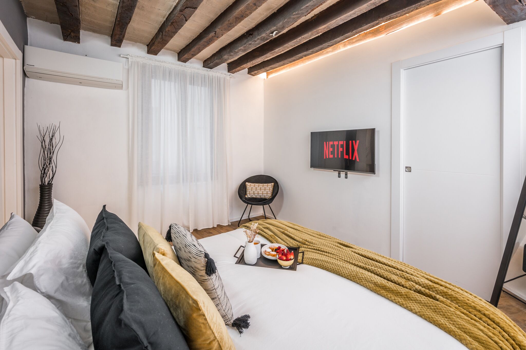 A modern bedroom with a bed, a bedside table with a tray of food, a chair, a TV mounted on the wall, and a window with curtains.