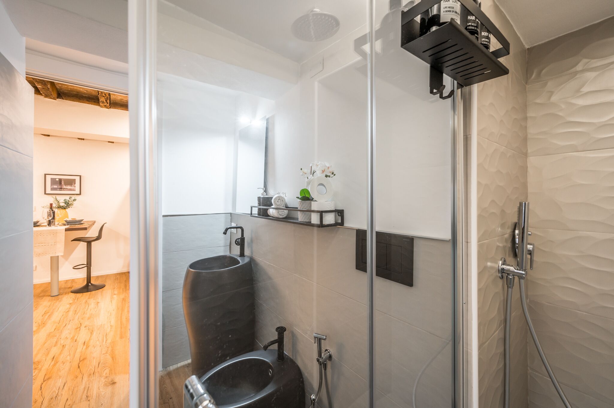 Un baño moderno con cabina de ducha de vidrio, un lavamanos negro y un estante pequeño con artículos de aseo.