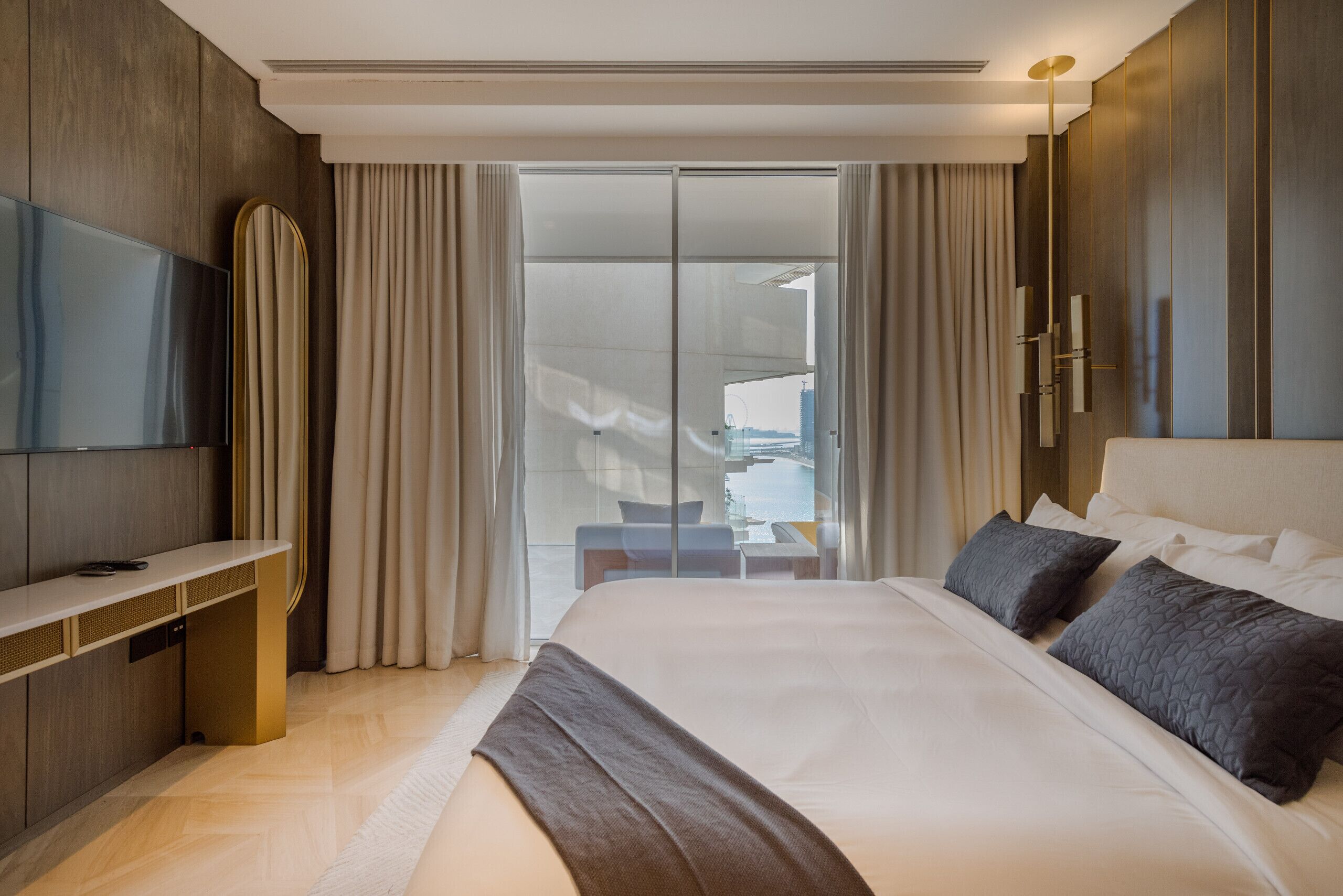 A modern hotel room with a large bed, a flat-screen TV, a desk, and a sliding glass door leading to a balcony.