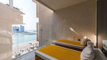 A modern hotel room with a large balcony, a bed with yellow bedding, and a view of a cityscape and water.