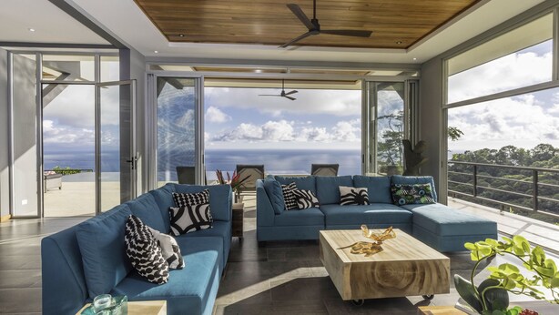 Ein modernes Wohnzimmer mit zwei blauen Sofas, einem Holztisch und einem großen Fenster mit Blick auf Himmel und Wolken.