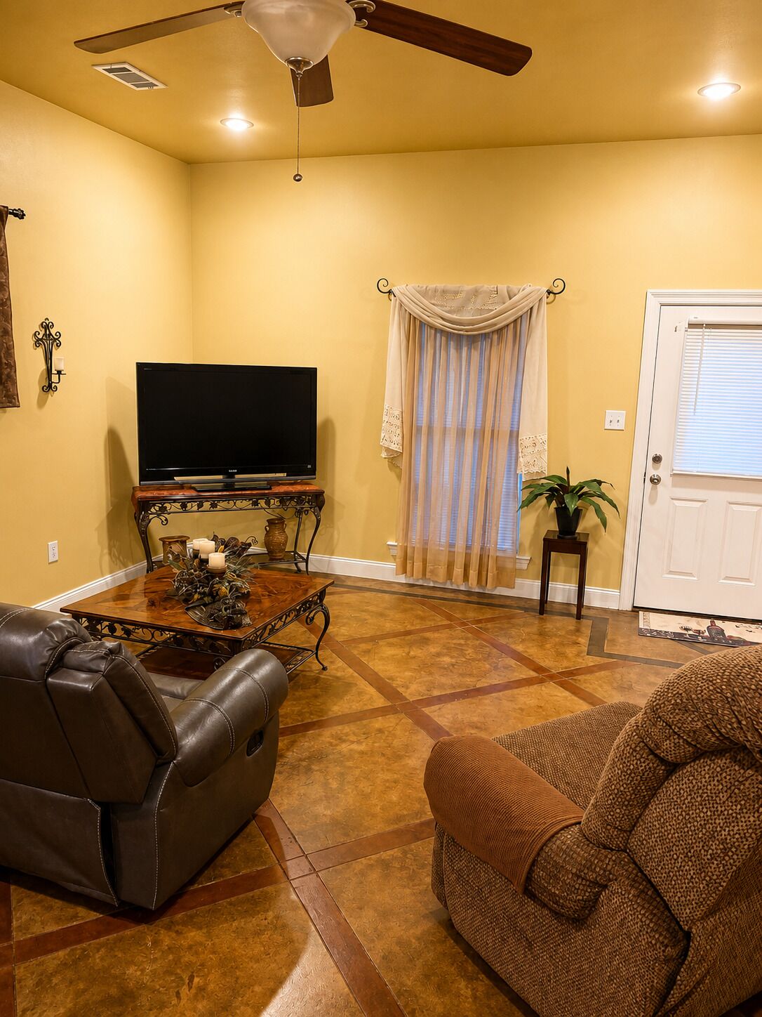 Una sala de estar con ventilador de techo, un televisor de pantalla plana sobre un soporte de madera, una mesa de comedor con velas y una puerta con ventana.