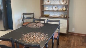 A dining area with a black table featuring a geometric pattern, surrounded by six chairs with brown cushions. A chandelier hangs from the ceiling, and a wooden cabinet with decorative plates and bowls is mounted on the wall. - Relax in  style! 3 BR ranch near downtown! FENCED yard! PETS welcome (Knoxville)