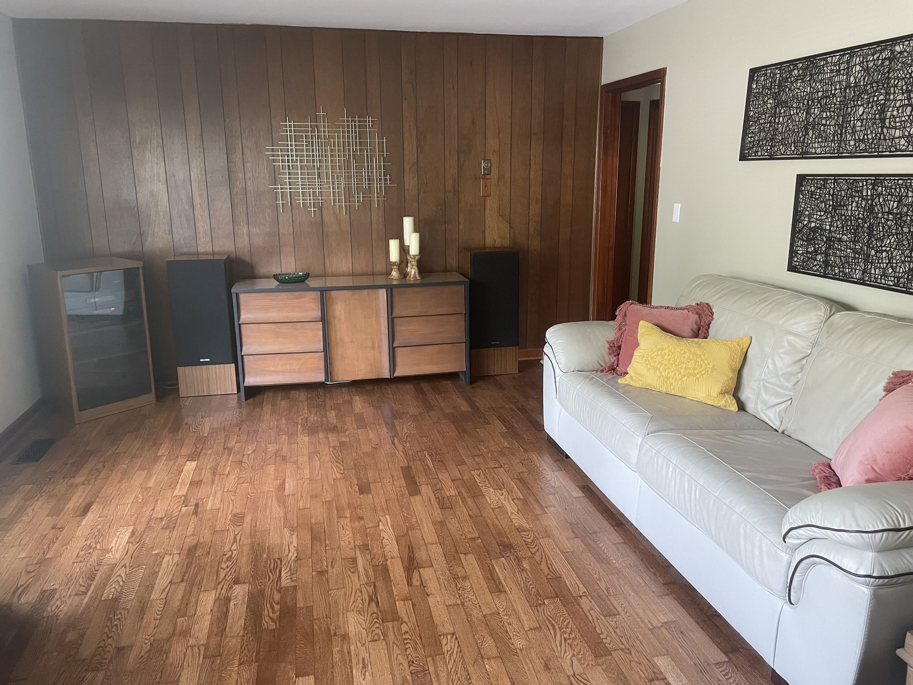 A living room with a wooden floor, a white sofa with cushions, a wooden cabinet with drawers, a speaker, and a wall-mounted light fixture.