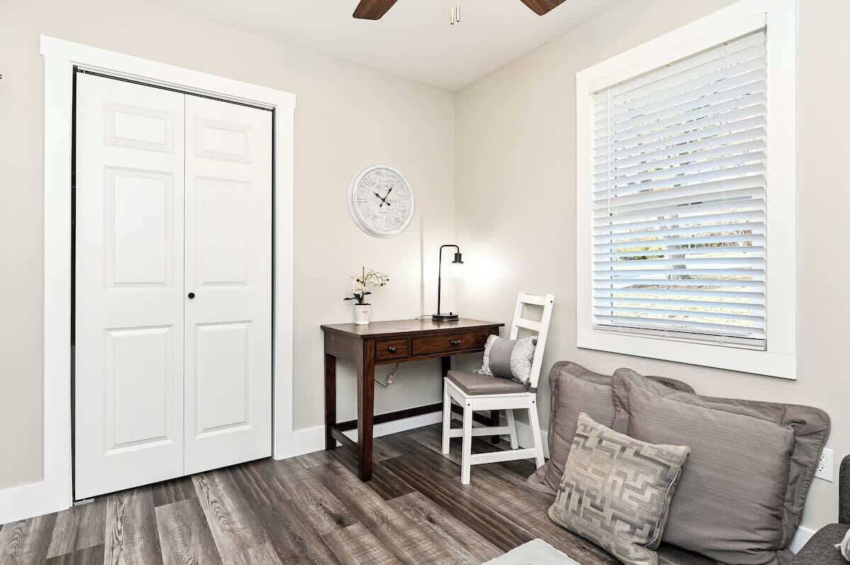 A room with a desk, chair, and a couch. There is a white door, a clock on the wall, and a window with blinds.