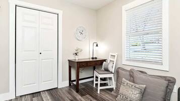 A room with a desk, chair, and a couch. There is a white door, a clock on the wall, and a window with blinds.