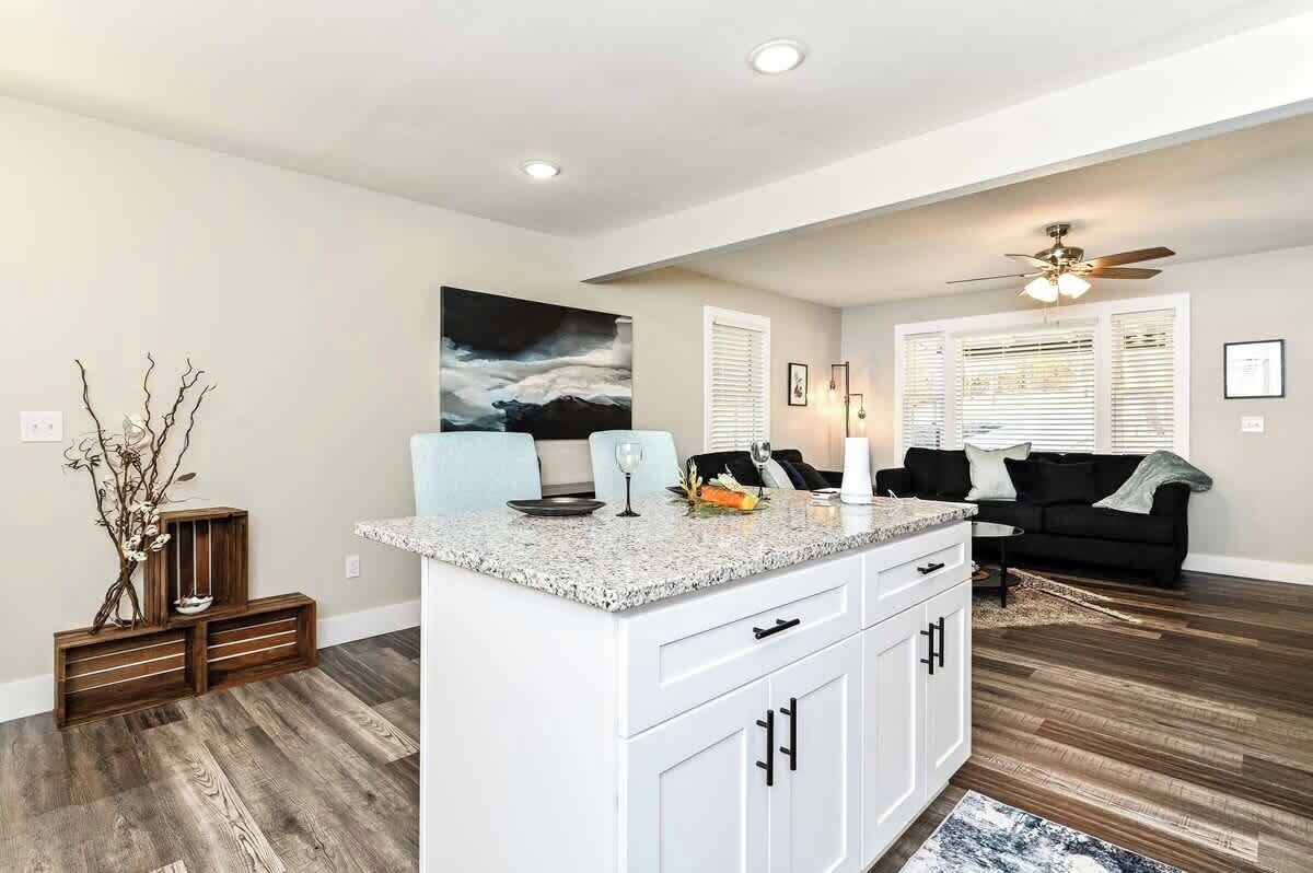 A modern kitchen with white cabinetry, a granite countertop, and a built-in island. There is a black couch in the adjacent living area, and a ceiling fan above.