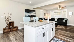 A modern kitchen with white cabinetry, a granite countertop, and a built-in island. There is a black couch in the adjacent living area, and a ceiling fan above. - Greenville Gateway: Elegant Urban Retreat (Greenville)