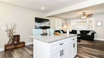 A modern kitchen with white cabinetry, a granite countertop, and a built-in island. There is a black couch in the adjacent living area, and a ceiling fan above.