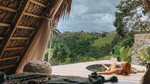 Una habitación con techo de paja, una cama, una persona acostada en la cama y vistas a un paisaje verde exuberante.