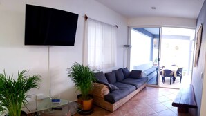 A living room with a sofa, two potted plants, a glass coffee table, and a flat-screen TV mounted on the wall.