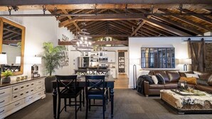 Een ruime woonkamer met een hoge plafond, zichtbare houten balken, een eettafel met stoelen, een leren bank en een grote spiegel.