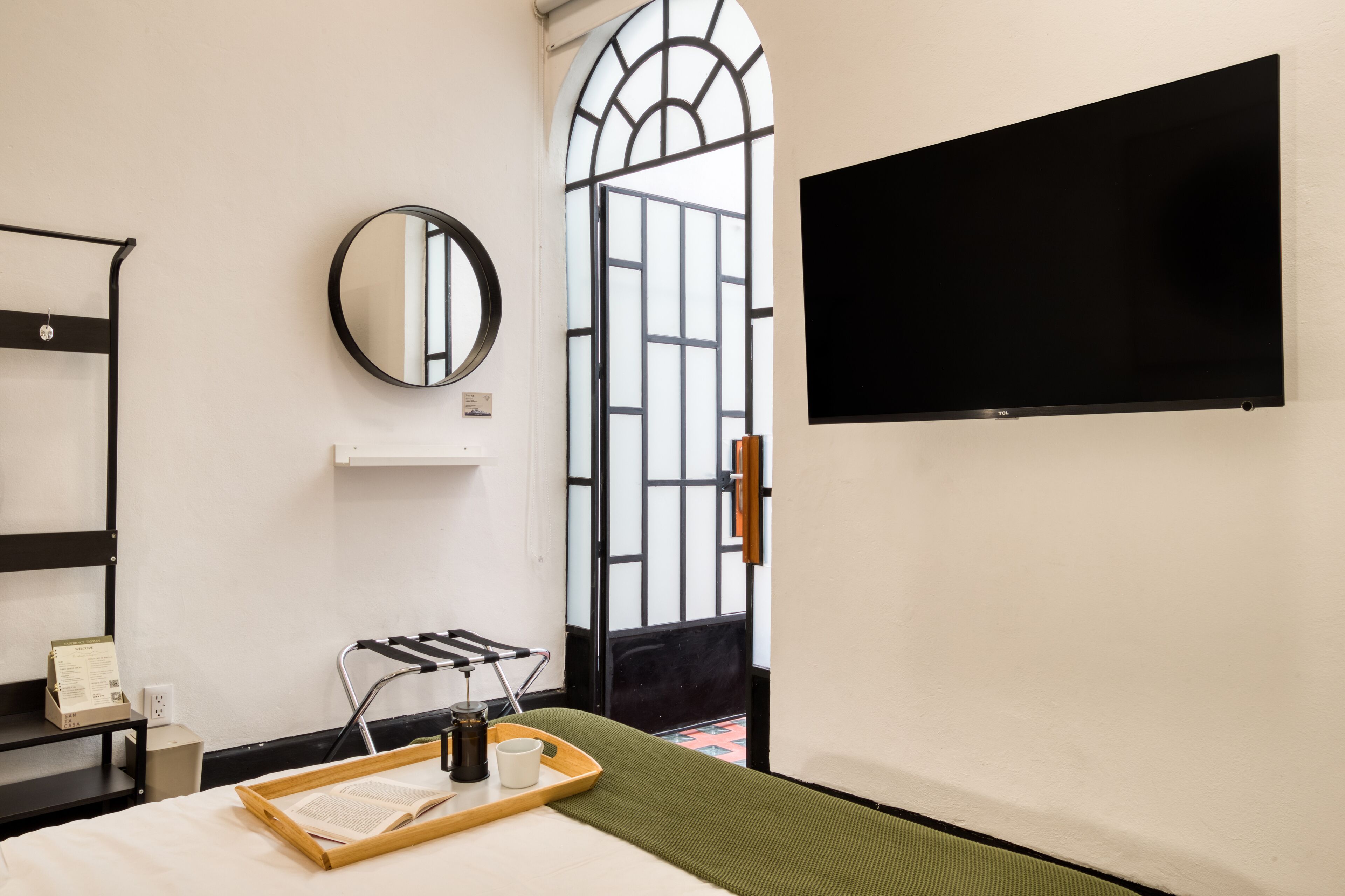 A hotel room with a bed, a TV, a mirror, a window with a view, and a tray with a book and a cup.