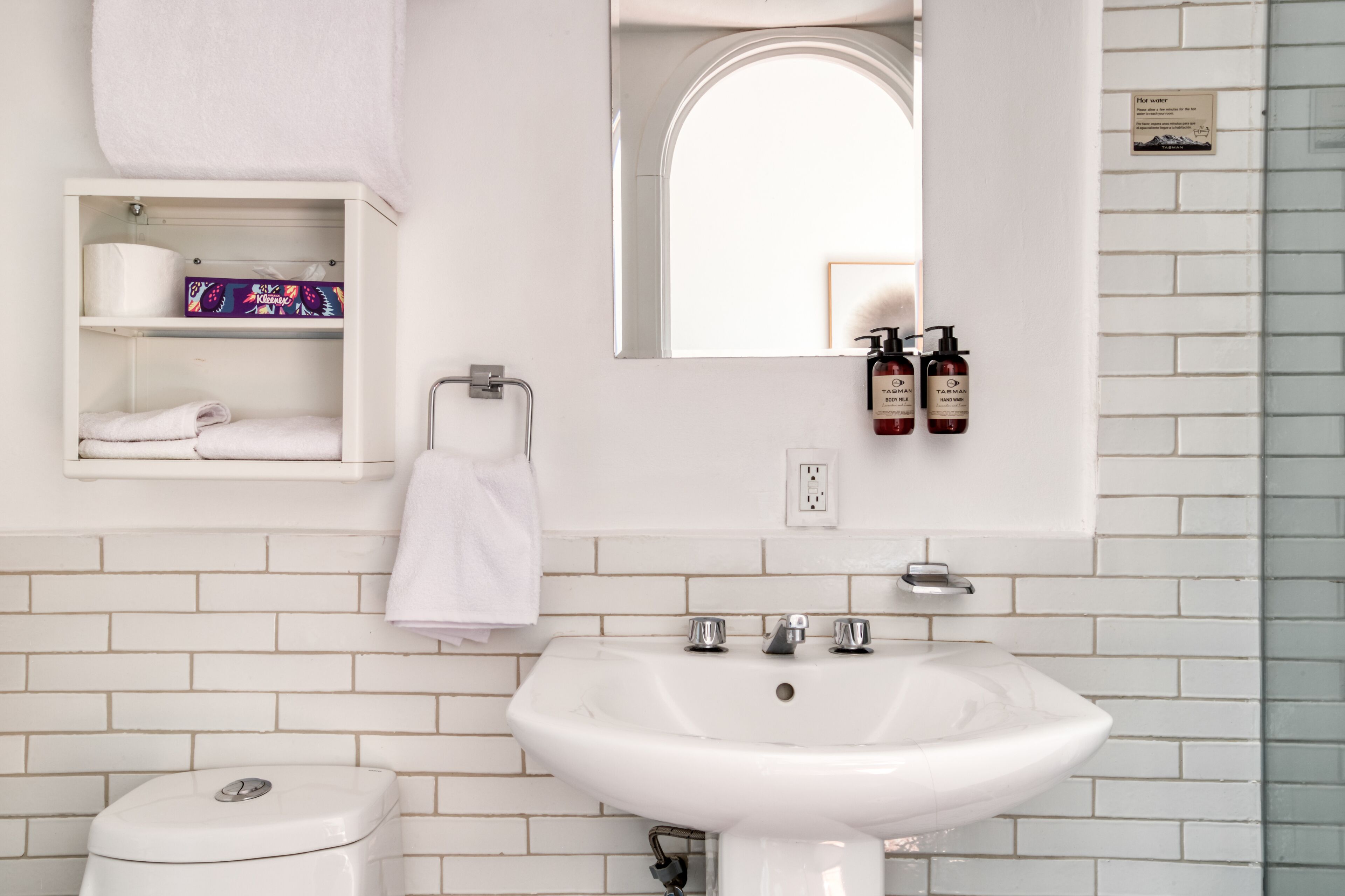 A bathroom with a white sink, a toilet, a shelf with toiletries, and a mirror.