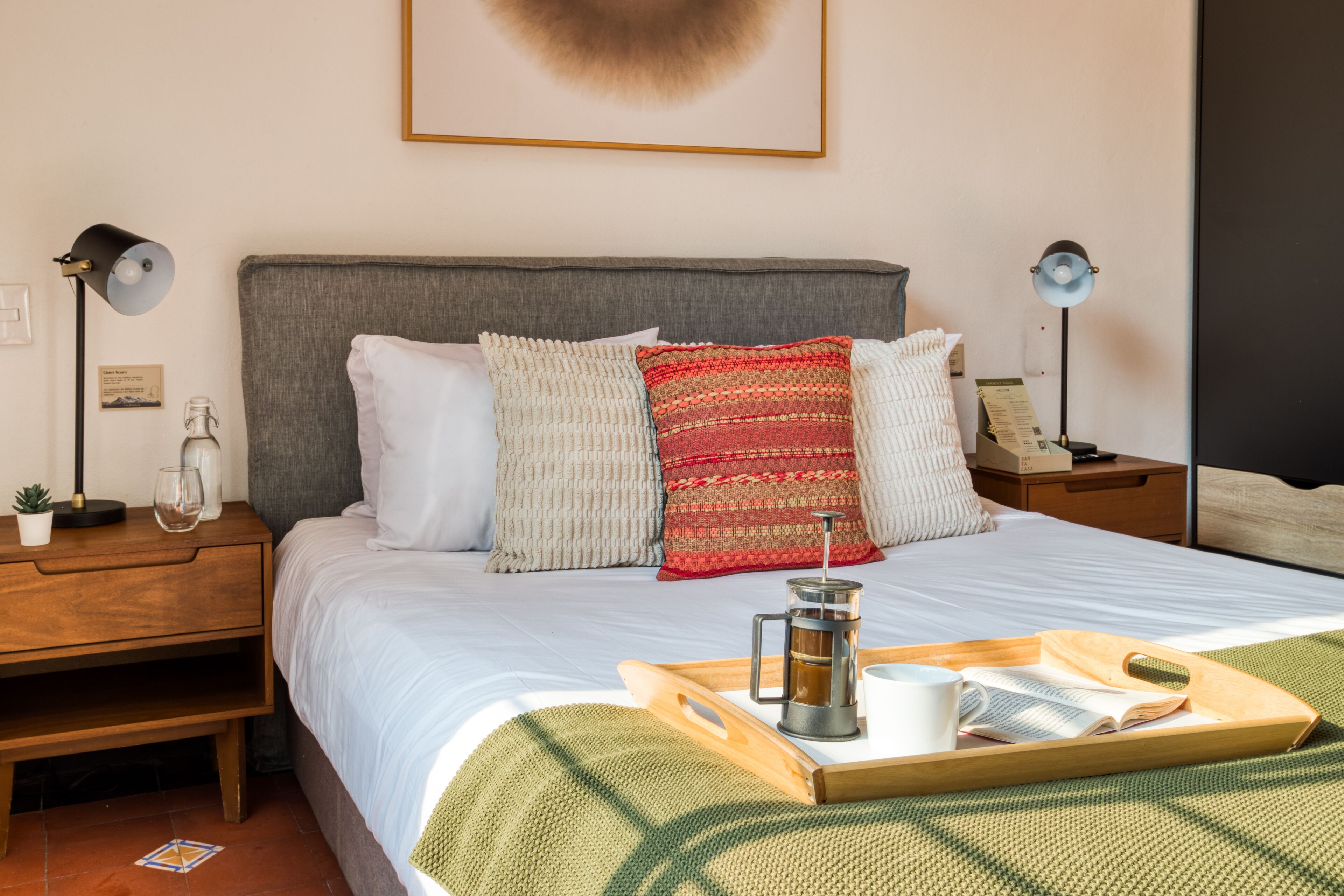 Een net opgemaakt bed met een dienblad vol ontbijtartikelen, een koffiezetapparaat en een boek op de nachtkastje.