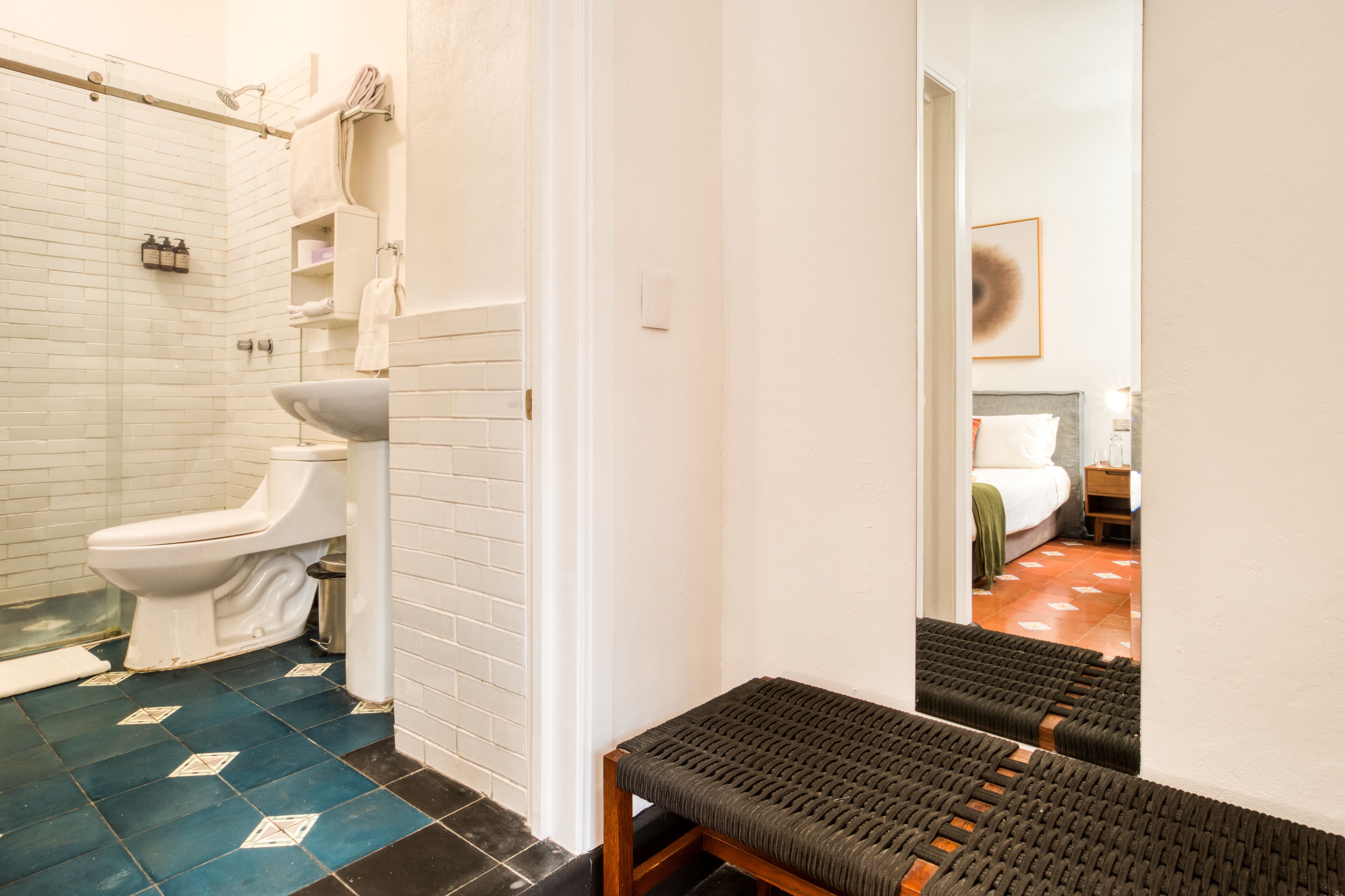 Een badkamer met toilet, wastafel en douche. Op de voorgrond staat een bankje en op de achtergrond een bed.