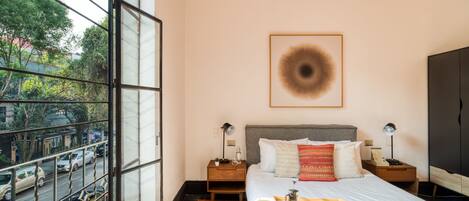 A bedroom with a large window, a bed with a green blanket, two bedside tables with lamps, and a framed picture on the wall.
