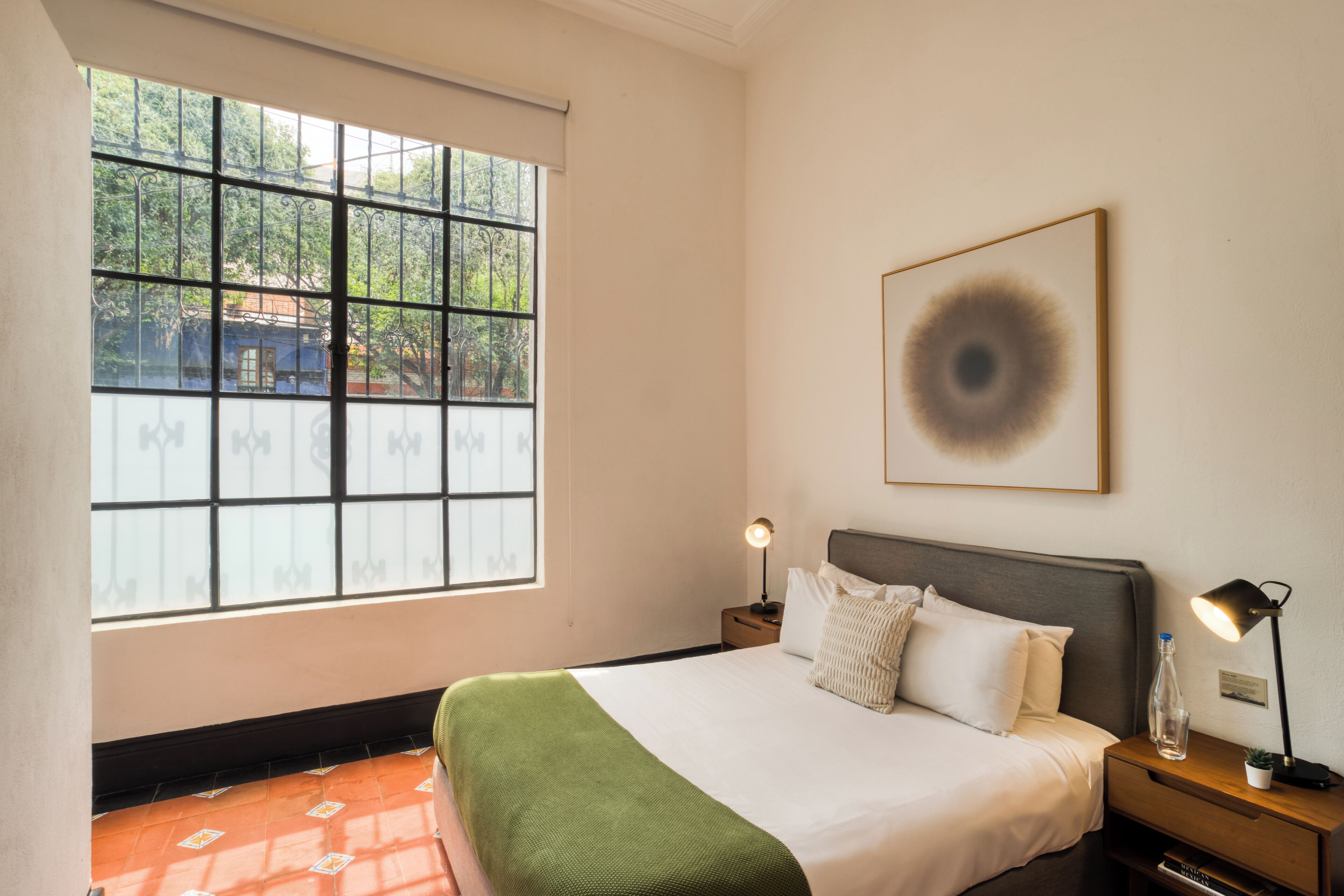 Une chambre à coucher bien rangée, avec une grande fenêtre, un lit recouvert d’un plaid vert, deux tables de chevet avec des lampes et une œuvre d’art encadrée accrochée au mur.