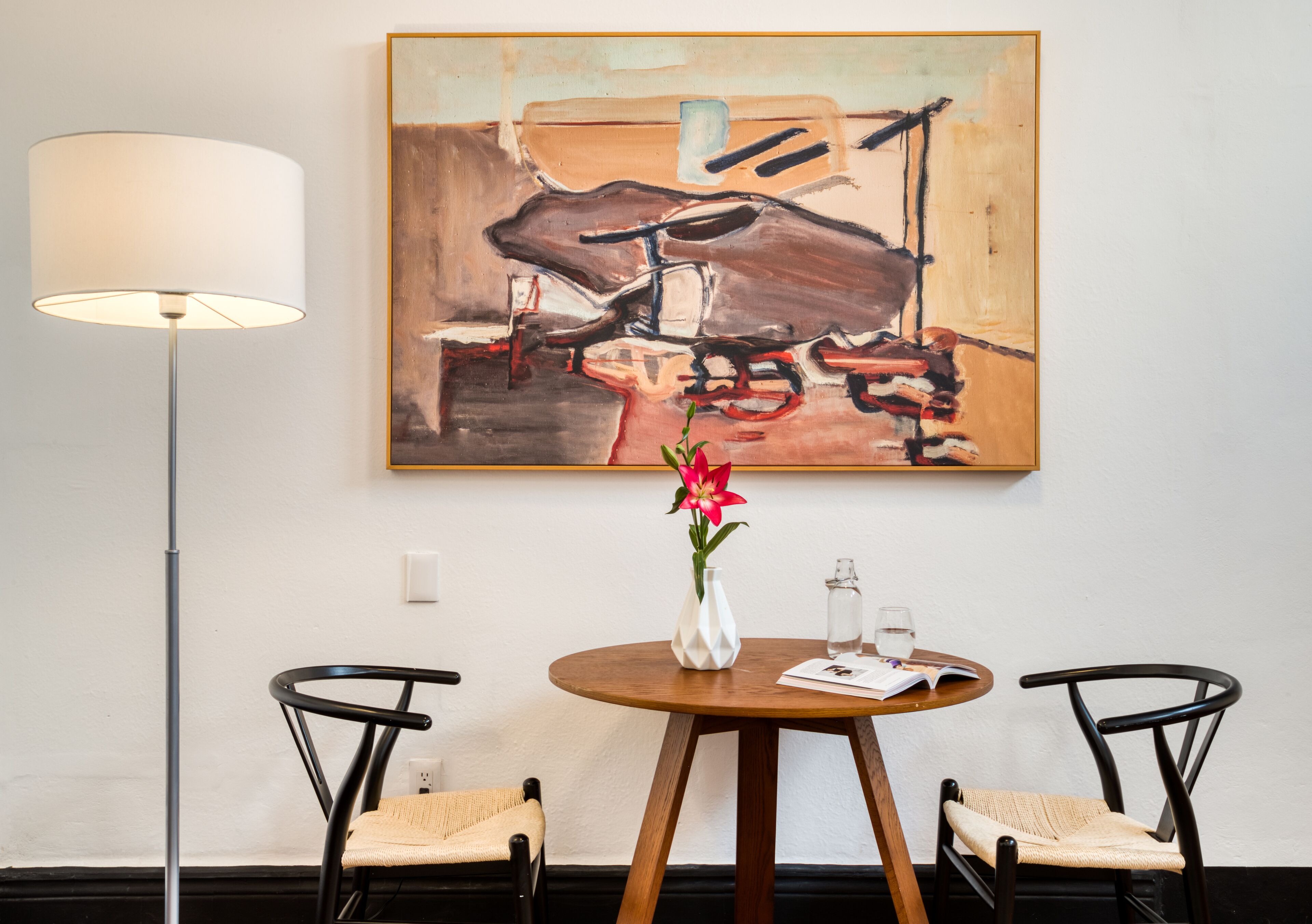 Een kamer met een ronde houten tafel, twee zwarte stoelen, een witte vaas met bloemen, een lamp en een schilderij aan de muur.