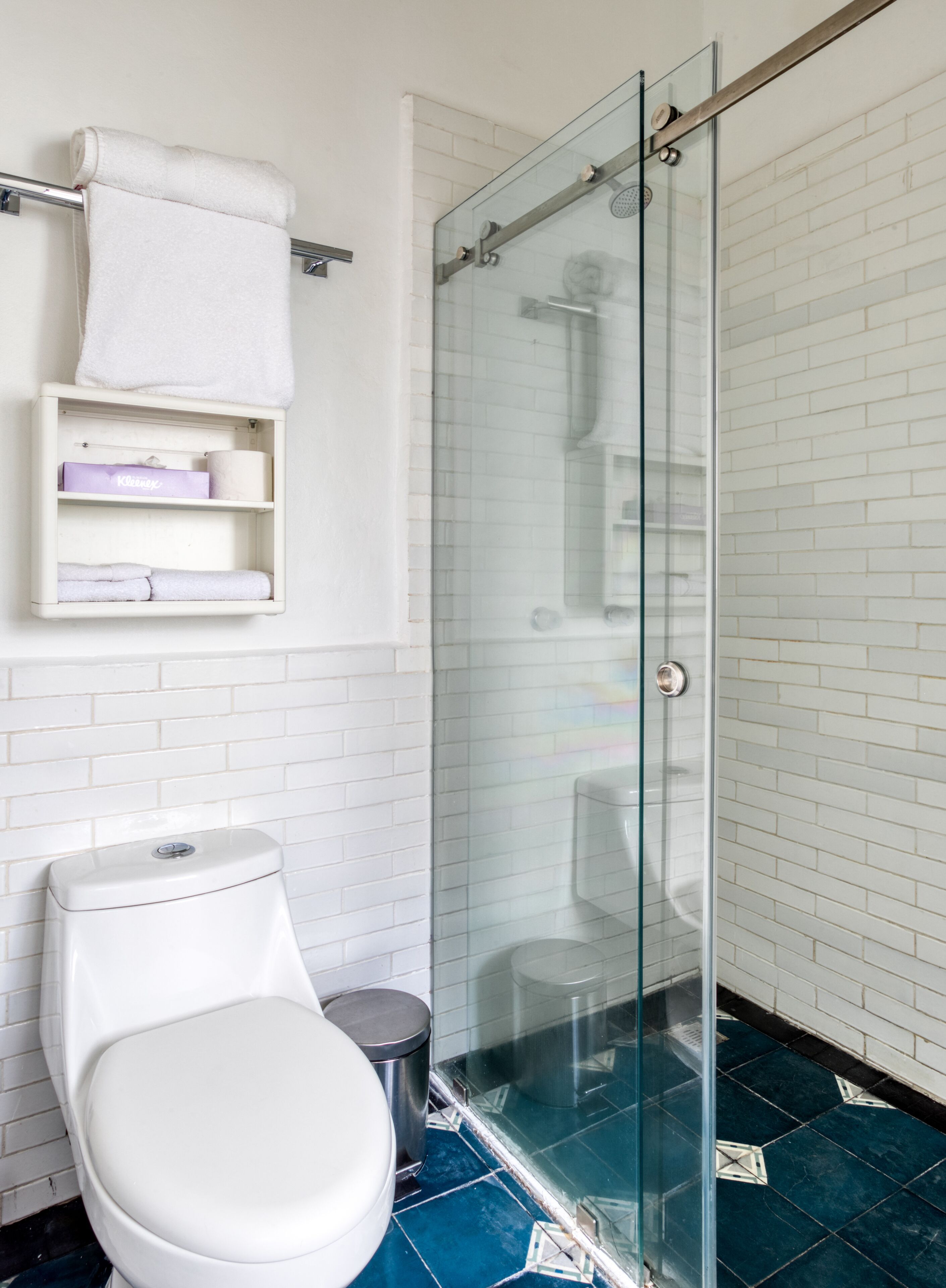 Un baño moderno con cabina de ducha de vidrio, paredes cubiertas de azulejos blancos, un inodoro blanco y un estante pequeño con artículos de aseo.
