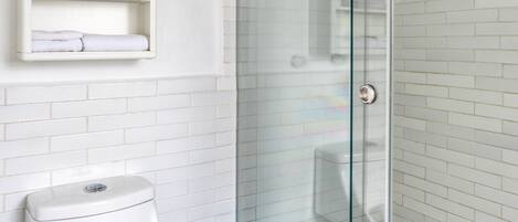 Un baño moderno con cabina de ducha de vidrio, paredes cubiertas de azulejos blancos, un inodoro blanco y un estante pequeño con artículos de aseo.
