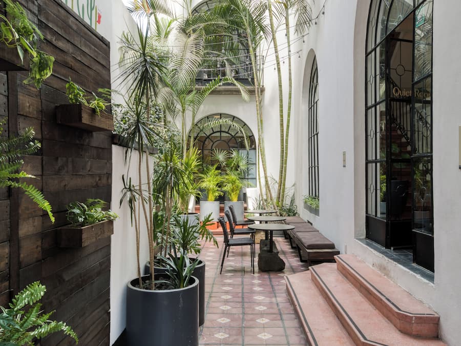 Un patio estrecho con paredes de madera, plantas en macetas, una zona de estar con una mesa y sillas, y una escalera que lleva a un nivel superior.