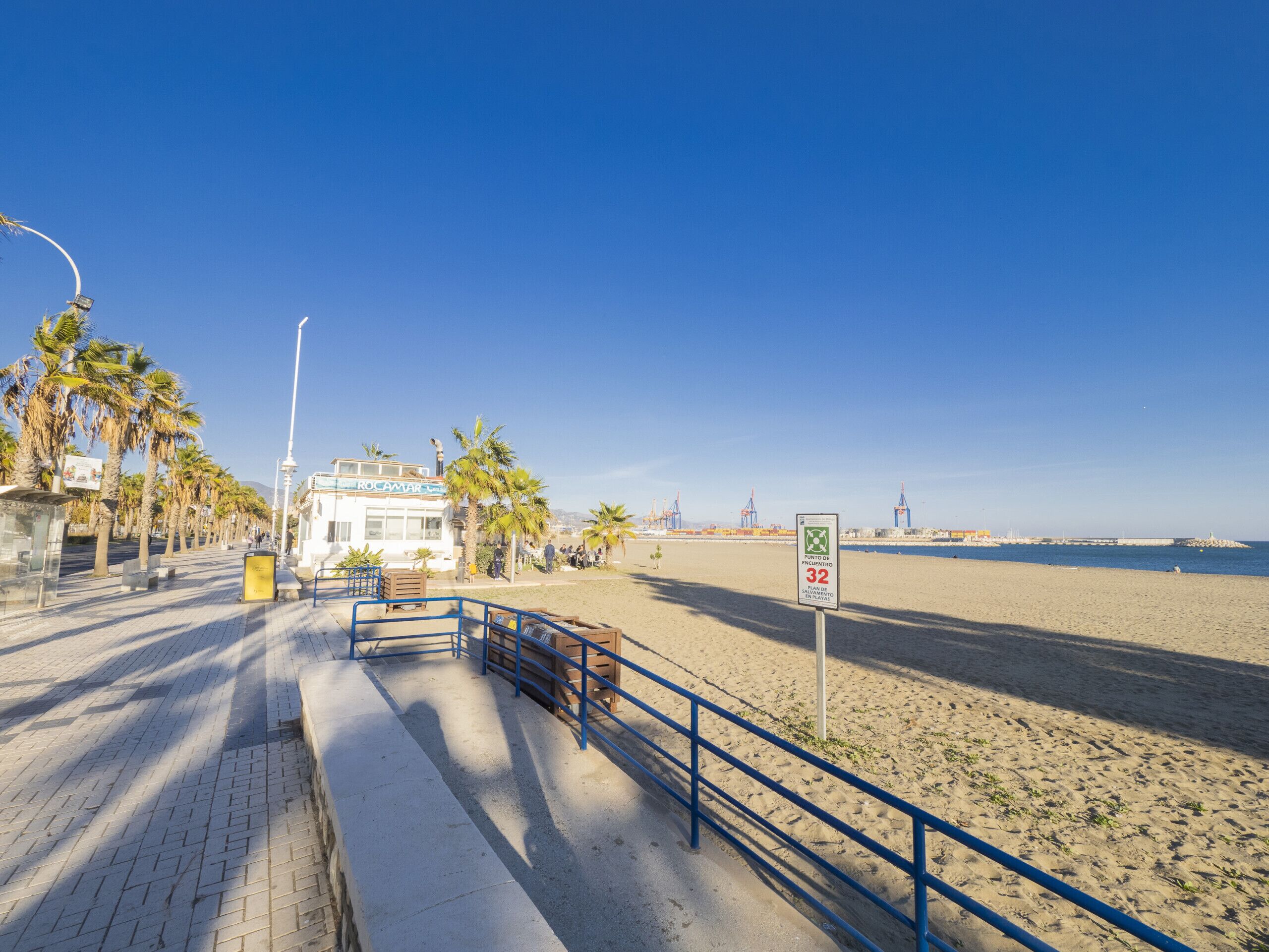 Een promenade aan het strand, met een heldere lucht, palmbomen, een blauwe reling en een bord dat een snelheidslimiet van 32 km/u aangeeft.