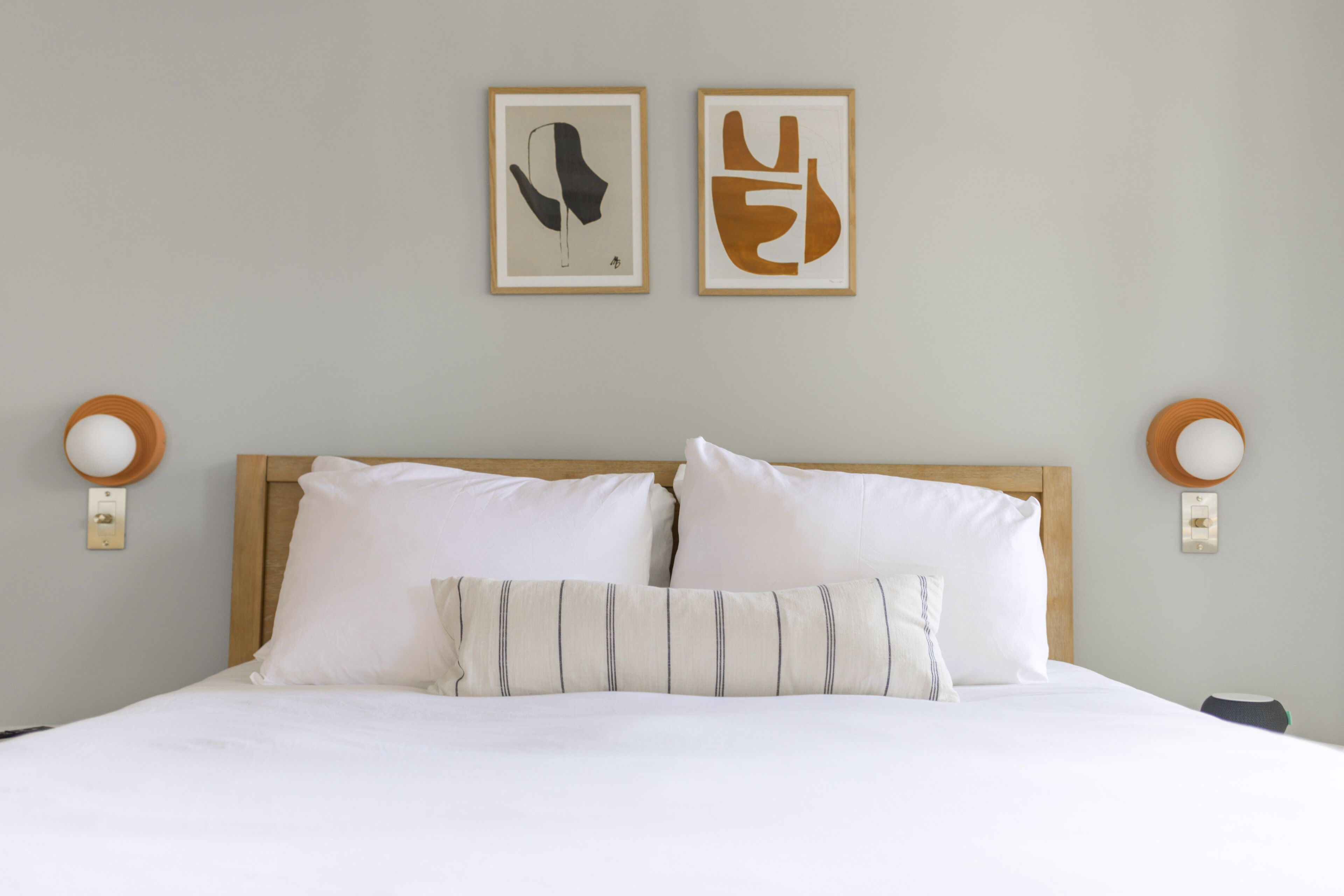 A neatly made bed with white linens and striped pillows, a wooden headboard, and two wall-mounted light fixtures.