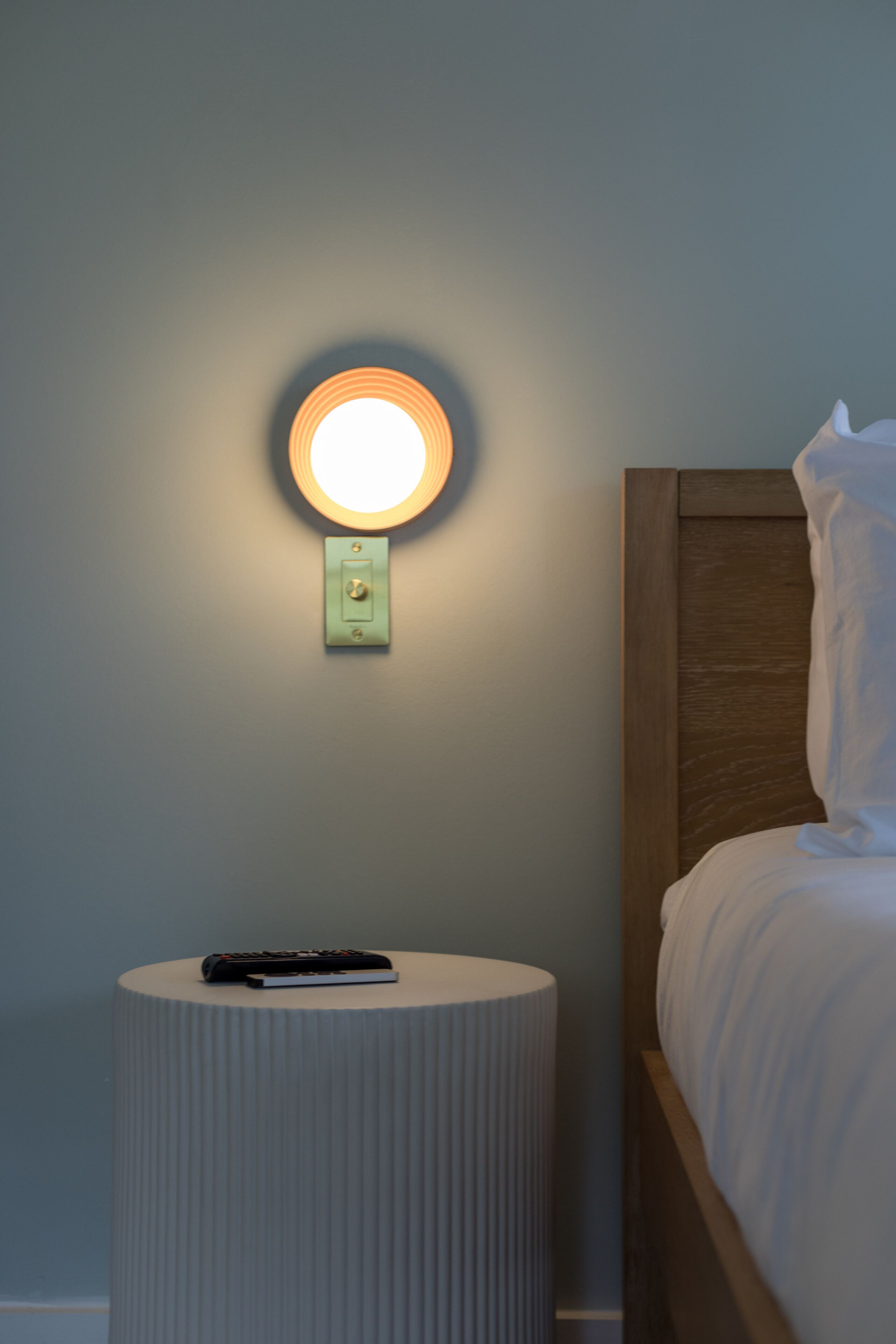 A neatly made bed with a wooden headboard, a round wall-mounted light fixture, and a small table with a remote control.