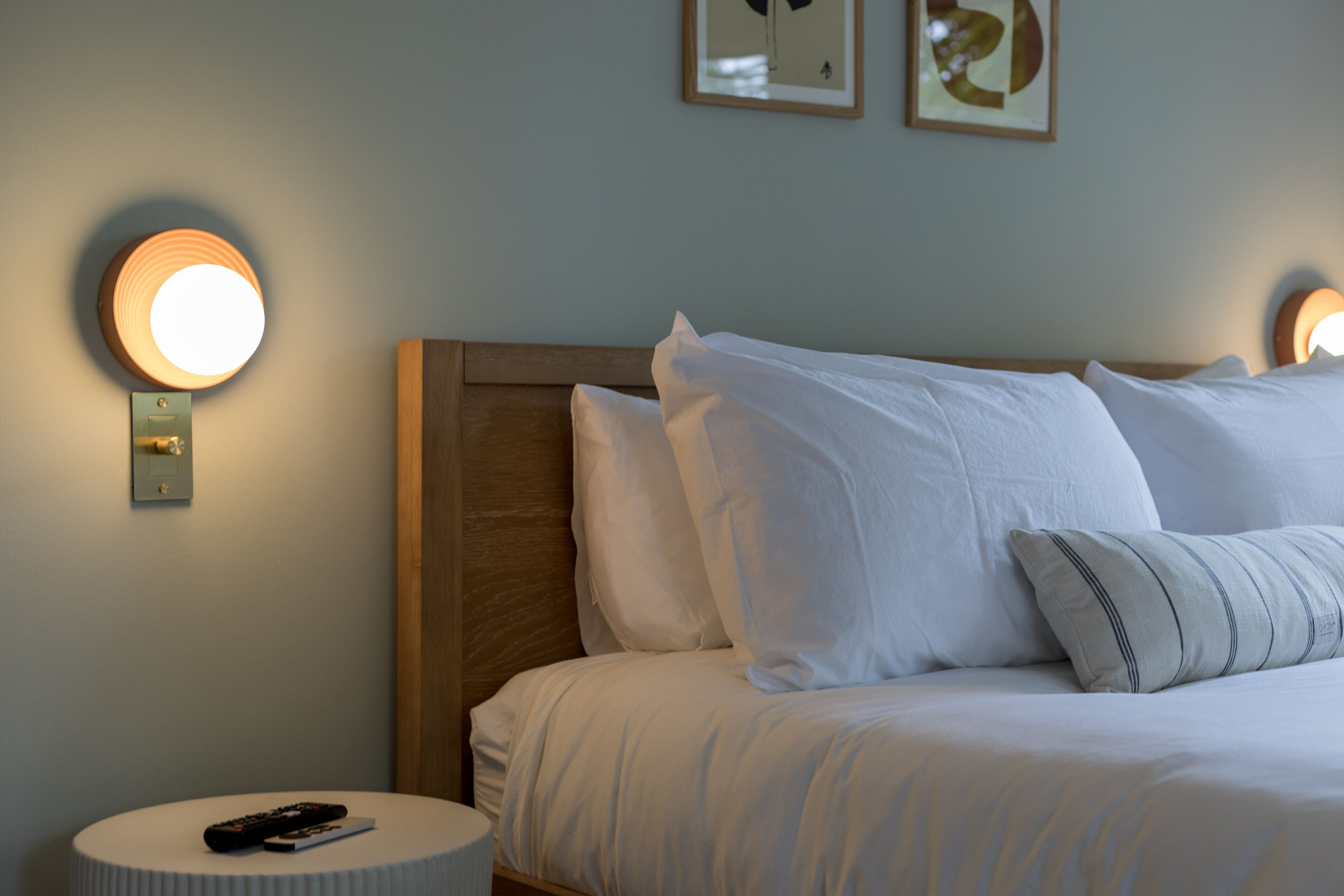 Cama bem arrumada com lençóis e almofadas brancos, mesa de cabeceira redonda com um controle remoto e luminárias fixadas na parede.